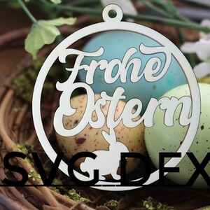 Pode incluir: Ornamento circular de madeira branco com o texto "Frohe Ostern" em uma fonte decorativa. O ornamento está sentado em um ninho de galhos com ovos de Páscoa salpicados.