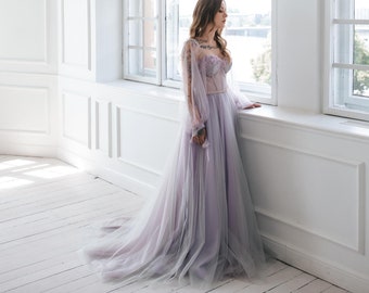 May include: A woman in a light purple tulle gown with a fitted bodice and long sleeves stands by a window in a white room.