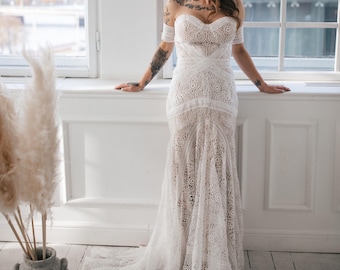 Puede incluir: Una mujer con un vestido de novia de encaje blanco con escote de corazón y tirantes de hombros descubiertos. Está de pie frente a una ventana con vista a la ciudad.