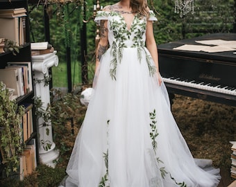 May include: A white wedding dress with a tulle skirt and a V-neckline, adorned with green leaf embroidery. The dress is worn in a greenhouse setting with a piano and chandeliers.