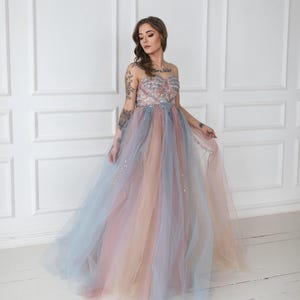 Peut inclure: Une femme portant une robe en tulle sans bretelles, multicolores, avec un corsage perlé. La robe présente un dégradé de couleurs rose, bleu et pêche.
