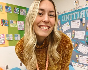 Peut inclure: Une femme aux longs cheveux blonds porte un pull moutarde et un jean bleu. Elle se tient dans une salle de classe aux murs colorés et une pancarte qui dit "Écriture".