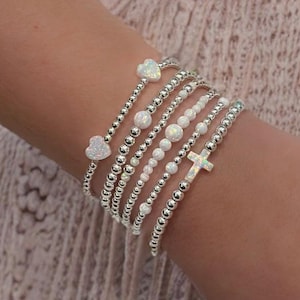 May include: A stack of silver beaded bracelets with opal heart and cross charms.