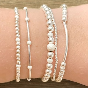May include: A set of five silver bracelets, including a beaded bracelet with textured beads, a beaded bracelet with smooth beads, a thin silver bangle, a thicker silver bangle, and a beaded bracelet with small beads.