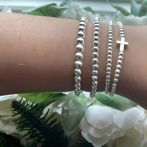 May include: Three silver beaded bracelets stacked on a wrist. The top bracelet has a small silver cross charm.