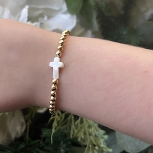 May include: A gold beaded bracelet with a white cross charm. The bracelet is on a person's wrist.