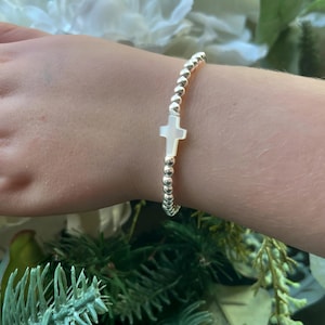 May include: A silver beaded bracelet with a white cross charm. The bracelet is on a person's wrist.