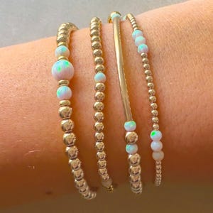 May include: Four gold bracelets with white opal beads. The bracelets are stacked on a person's wrist.