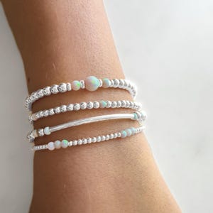 May include: A set of four silver bracelets with small white opal beads. The bracelets are stacked on a person's wrist.