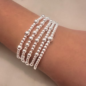 May include: A set of six silver beaded bracelets stacked on a person's wrist. The bracelets are made of small, round beads and are all the same size.
