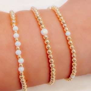 May include: Three gold beaded bracelets with white opal accents. The bracelets are stacked on a person's wrist.