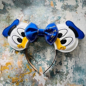 May include: A Donald Duck-themed headband with white, yellow, and blue features. The headband has two large, cartoonish Donald Duck faces, with a blue bow in the centre. The bow has gold text that reads "Donald Duck".