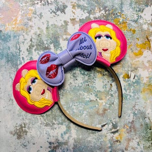 May include: A pair of pink mouse ears featuring a cartoon character with blonde hair and a bow with the text "All about Moi!" and red lips. The ears are attached to a brown headband.