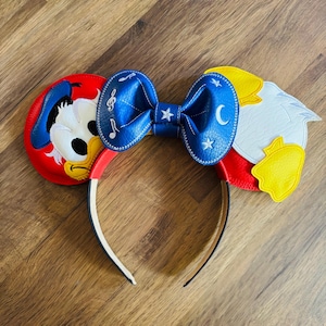 May include: A Disney-themed headband featuring Donald Duck on one ear and a stylized eagle on the other. The headband has a blue bow adorned with musical notes, stars, and a crescent moon. The ears are red, yellow, and white.
