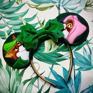 May include: A pair of mouse ears featuring a green bow with arrow details and two circular ear designs. One ear shows a fox character in a green hat, and the other shows a pink-clad character. The headband is tan.