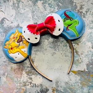 May include: A whimsical headband featuring mouse ears with cartoon character designs. The ears are light blue with embroidered images of a crowned lion and a snake. A red bow with white polka dots sits between the ears. The headband is brown.