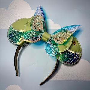 May include: Green and blue iridescent mouse ears with a swirling pattern. The ears are attached to a brown headband.
