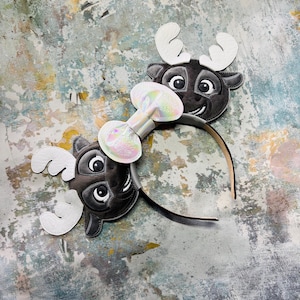 May include: Two headbands with reindeer designs. Each headband features a dark grey reindeer face with white antlers and a smiling expression. A shimmering bow is attached between the reindeer faces.