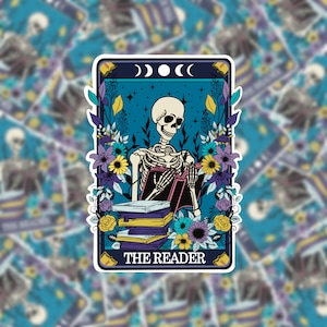 May include: A tarot card illustration featuring a skeleton reading a book. The card is blue with a crescent moon design and the text "THE READER" at the bottom. The skeleton is surrounded by colourful flowers.