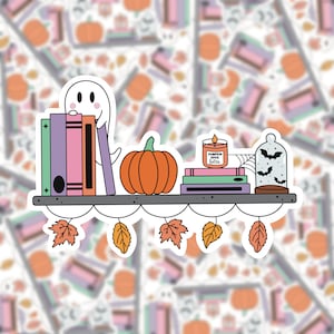 May include: A white ghost with a friendly face peeks out from behind a stack of colorful books on a shelf. A pumpkin, a candle that says "Pumpkin Spice Latte", and a glass cloche with bats inside complete the autumnal scene.  Fall leaves hang from the shelf.