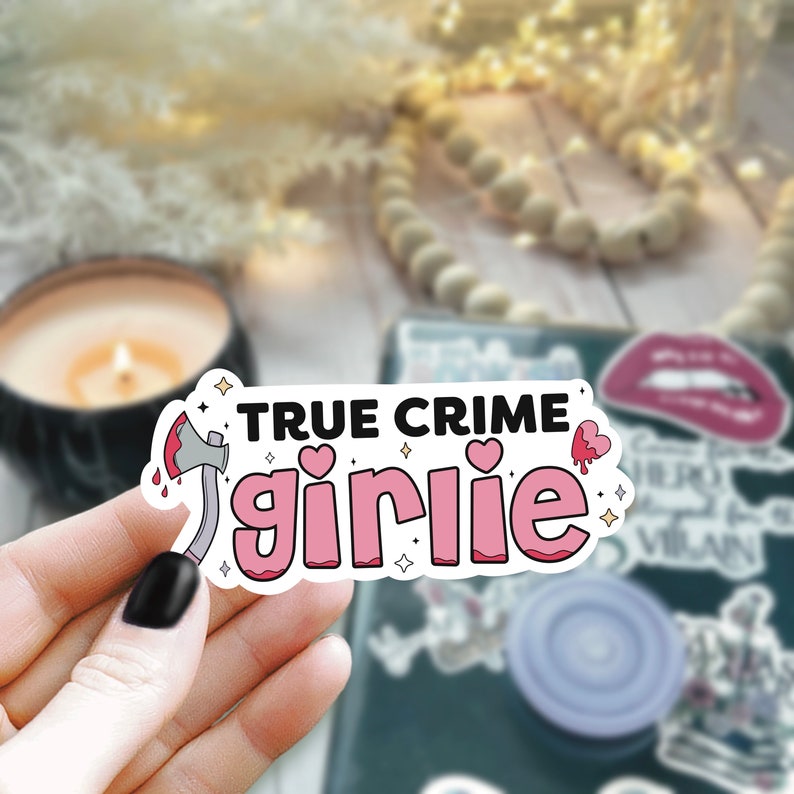 May include: A pink and white sticker with the text "TRUE CRIME girlie" and a cartoon axe with a red handle and a dripping red heart. The sticker has a white outline and a glittery background.