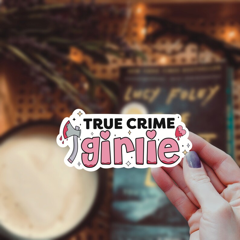 May include: A pink and white sticker with the text "TRUE CRIME girlie" in pink letters. The sticker features a cartoon axe with a red handle and dripping blood. There are also small pink hearts and stars around the text.