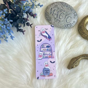 May include: A purple bookmark with a spooky theme featuring ghosts reading books. The bookmark has three scenes: a ghost reading in a coffin, a ghost reading in a bookshelf, and a ghost reading on a tombstone. The text on the tombstone reads "Reading in Peace".