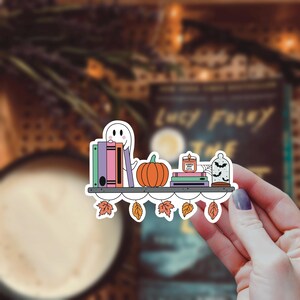 May include: A white ghost with a friendly face sits on a bookshelf with a stack of colorful books, a pumpkin, a candle that says "Pumpkin Spice Latte", and a glass cloche with bats. The bookshelf has a white scalloped edge and is decorated with fall leaves.