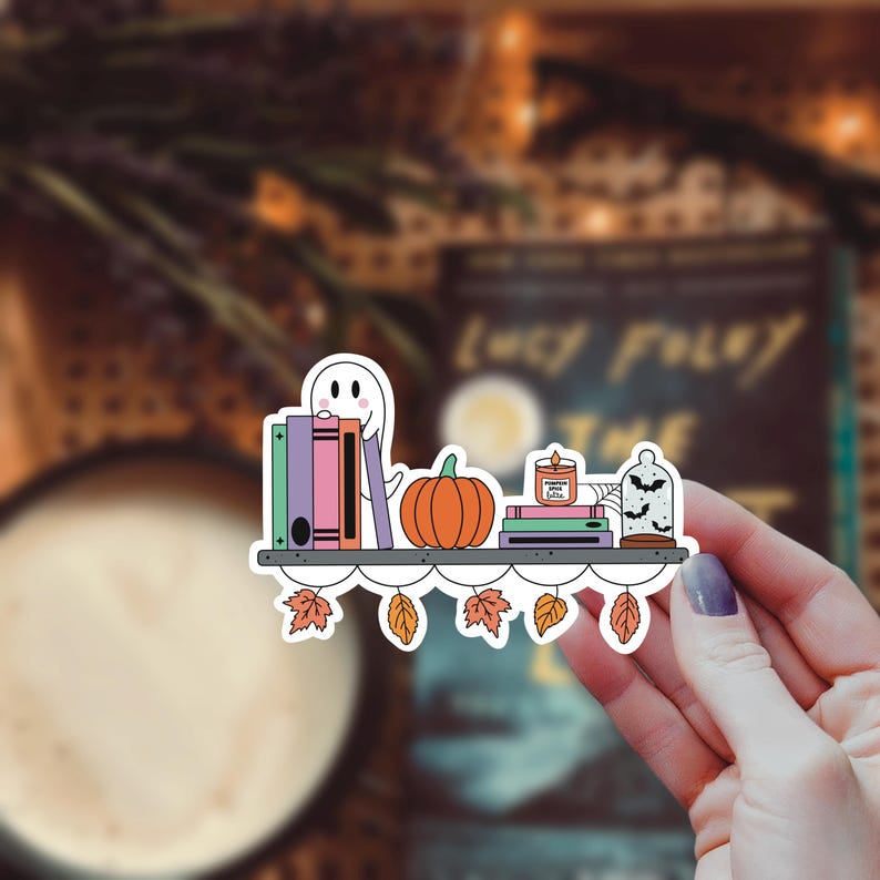 May include: A white ghost with a friendly face sits on a bookshelf with a stack of colorful books, a pumpkin, a candle that says "Pumpkin Spice Latte", and a glass cloche with bats. The bookshelf has a white scalloped edge and is decorated with fall leaves.