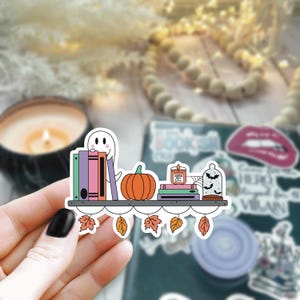 May include: A sticker featuring a bookshelf with a ghost, pumpkin, books, a candle that says "Pumpkin Spice Latte", and a glass dome with bats. The shelf is decorated with fall leaves.