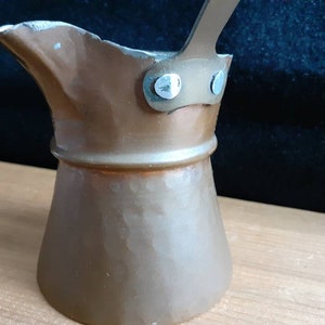 Puede incluir: Una cafetera de cobre martillado con un asa de metal y dos remaches. La cafetera tiene un cuerpo redondeado y un pico.