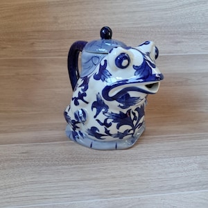 May include: A blue and white ceramic teapot shaped like a frog. The teapot has a floral design and a spout that looks like the frog's mouth.