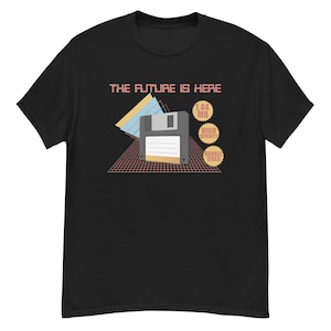 Puede incluir: Camiseta negra con un diseño retro que presenta un gráfico de disquete. El texto "THE FUTURE IS HERE" está encima del gráfico, con "1.44 MB", "HIGH DENSITY" y "DOUBLE SIDED" en un círculo a la derecha.