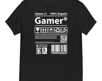 Op de afbeelding: Zwart t-shirt met het woord "Gamer" in witte, vette letters. Het ontwerp bevat een grafisch label met pictogrammen en tekst, waaronder "100% Organic" en een barcode. De ESRB-classificatie is "E 3+".