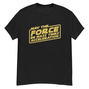 Puede incluir: Camiseta negra con un diseño rectangular dorado. El diseño incluye el texto "MAY THE FORCE BE MASS TIMES ACCELERATION" en una fuente estilizada. La camiseta está hecha de un material suave y tiene mangas cortas.