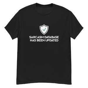 Op de afbeelding: Zwart T-shirt met de witte tekst "SARCASM DATABASE HAS BEEN UPDATED" en een wit schild-icoon met een groene vink. Het T-shirt heeft een ronde hals.