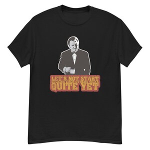 Può includere: T-shirt nera con un'immagine in scala di grigi di un uomo in giacca e cravatta che tiene una tazza. Sotto l'immagine, il testo recita "LET'S NOT START SUCKING EACH OTHERS DICKS QUITE YET" in un carattere invecchiato.
