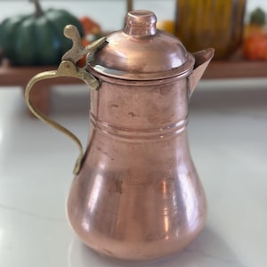 May include: A small copper pitcher with a brass handle and lid. The pitcher has a rounded body and a slightly flared spout.
