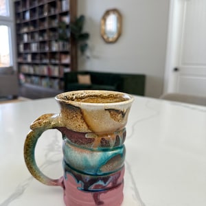 May include: A handmade ceramic mug with a unique, layered design. The mug features a handle and is painted with a blend of colors, including pink, teal, brown, and cream. The mug is sitting on a white surface.