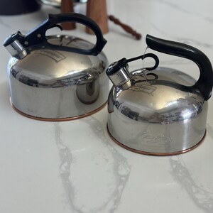 May include: Two stainless steel tea kettles with black handles and spouts. The kettles have a shiny, reflective surface and a copper-colored band around the base. The handles are arched, and the spouts have a whistle mechanism.