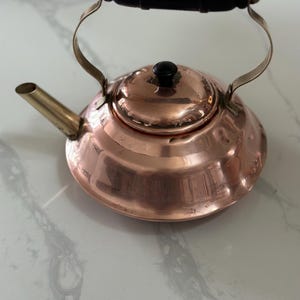 May include: A copper tea kettle with a wooden handle and a brass spout. The kettle has a shiny, polished finish.