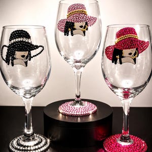 May include: Three clear glass wine glasses decorated with rhinestone embellishments. Each glass features a profile illustration of a person wearing a wide-brimmed hat. The bases are covered in black, light pink, and dark pink rhinestones.