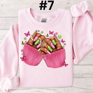 May include: Pink sweatshirt with a graphic of two hands with green and pink nail polish, rings, and butterflies. The text #7 is in the top center of the image.