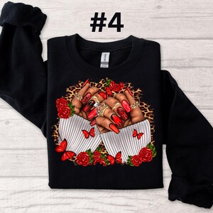May include: Black sweatshirt with a graphic print of two hands with red nail polish and rings, holding red roses and butterflies on a leopard print background. The text "#4" is printed on the top of the sweatshirt.