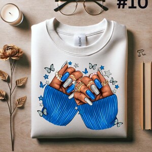 May include: White sweatshirt with a blue and white illustration of two hands with long, blue and silver painted nails. The hands are wearing rings and are surrounded by stars and butterflies.
