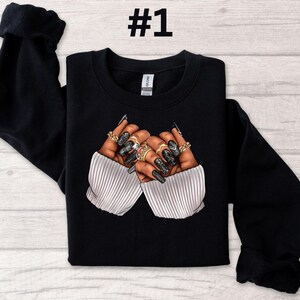 May include: Black sweatshirt with a graphic of two hands with long, black nails and gold rings. The hands are crossed over each other.