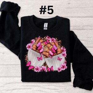May include: Black sweatshirt with a design of two hands with pink and white nail polish, rings, and butterflies. The design is surrounded by pink flowers and leopard print.