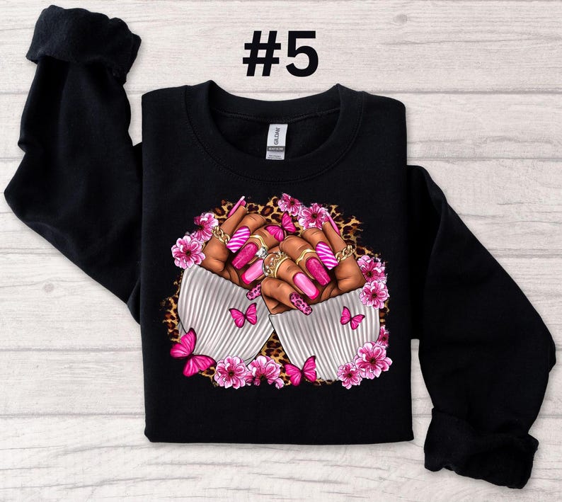 May include: Black sweatshirt with a design of two hands with pink and white nail polish, rings, and butterflies. The design is surrounded by pink flowers and leopard print.