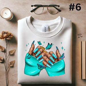 May include: White sweatshirt with a graphic of two hands with long, blue and white painted nails. The hands are holding a gold ring and are surrounded by blue butterflies.