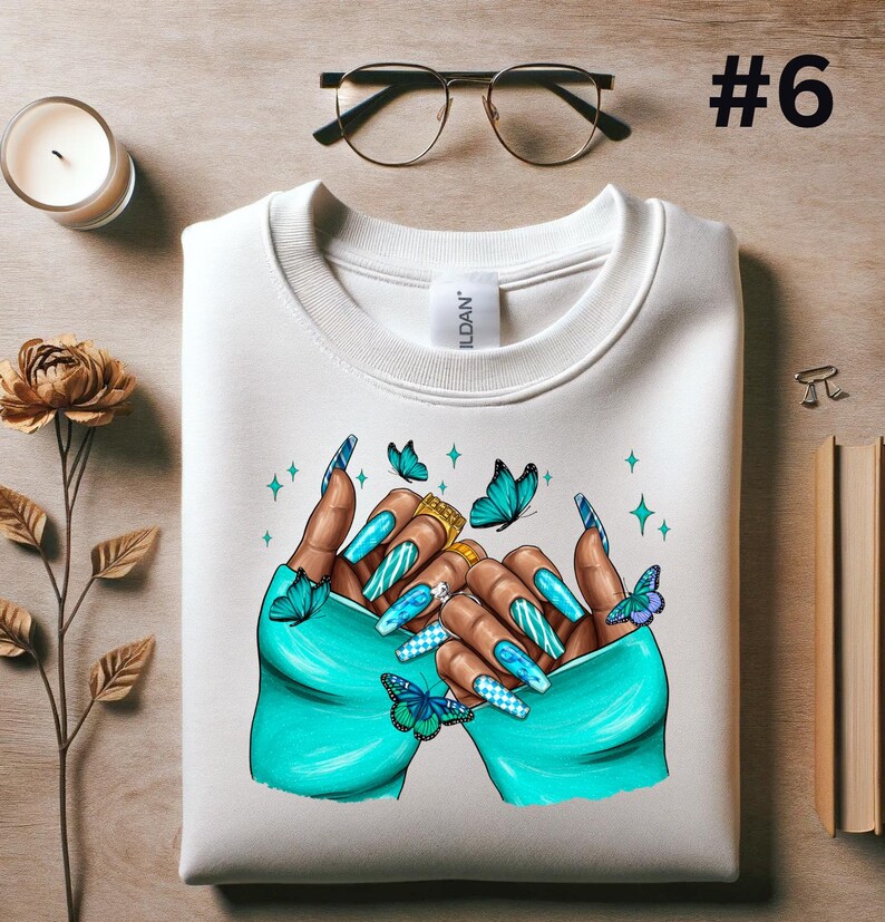 May include: White sweatshirt with a graphic of two hands with long, blue and white painted nails. The hands are holding a gold ring and are surrounded by blue butterflies.